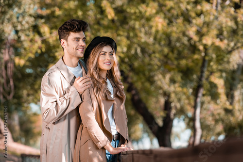 selective focus of man touching shoulders of woman in hat and trench coat in autumnal park