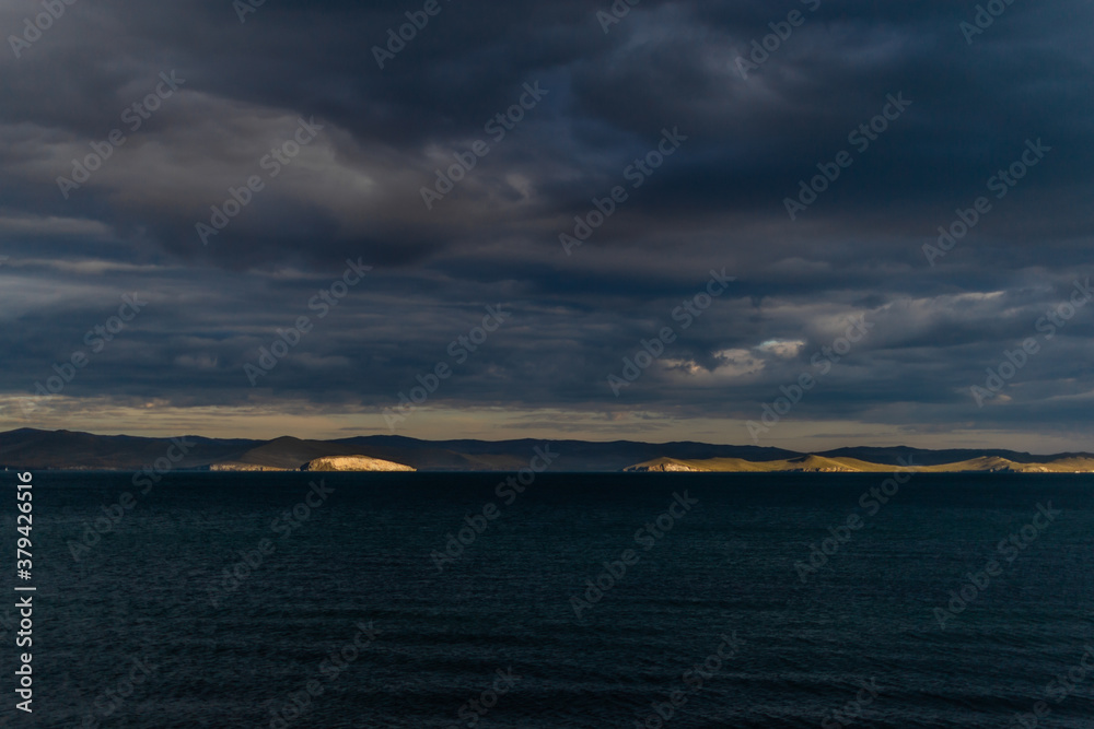 view of the clear calm undulating dark blue water of Lake Baikal, gold mountains in sunset light on the horizon, clouds, .ripples