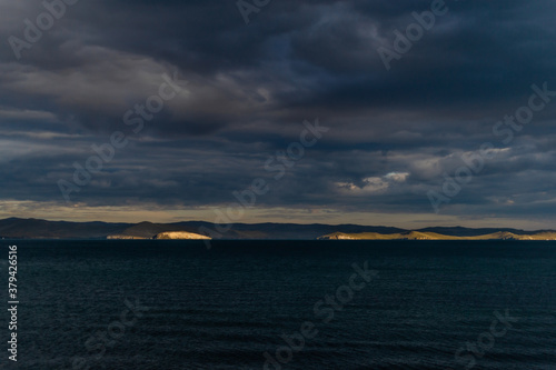 view of the clear calm undulating dark blue water of Lake Baikal, gold mountains in sunset light on the horizon, clouds, .ripples © SymbiosisArtmedia