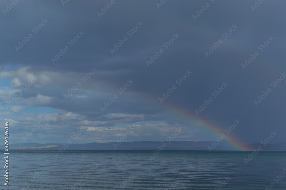 view of the clear calm undulating blue water of Lake Baikal, mountains on the horizon, colorful rainbow in sky