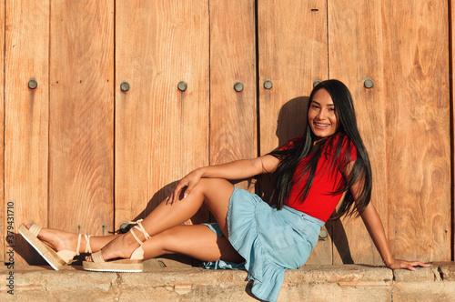 A young female on a summer day wearing a red shirt, bright blue skirt and beige espadrilles sandals posing sitting next to a big vintage door photo