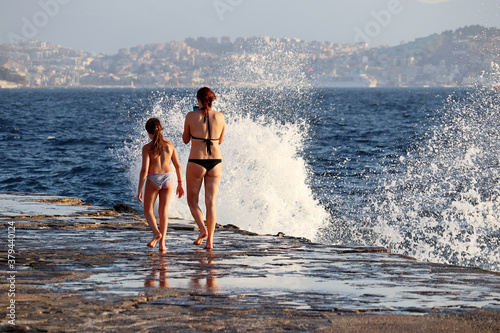 Woman in bikini walking with little girl on stone beach on surf waves background, rear view. Storm on a sea with water splashes, vacation and family leisure