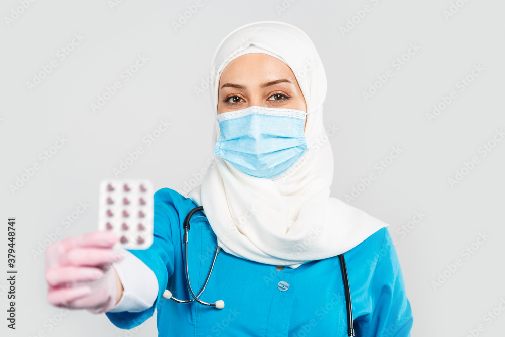 friendly Muslim doctor or nurse in a hijab, mask, gloves offering a pill to the patient on a gray background friendly Muslim doctor or nurse in a hijab, mask, gloves offering a pill to the patient on a gray background