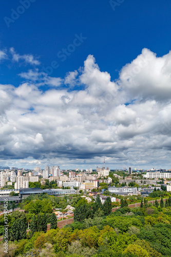 An urban landscape with a green park, residential areas and a TV tower against a bright blue sky with thickening clouds.