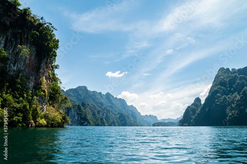 A magical view of the limestone cliffs in Cheow Lan Lake. A turquoise lake against the backdrop of huge rocks overgrown with tropical lush greenery and a blue sky with the clouds