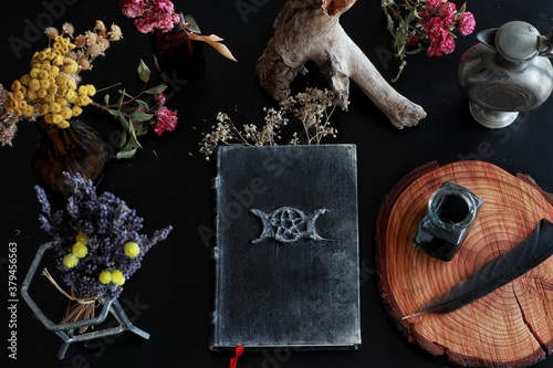 Flat lay of witch's altar space with hand made old looking book grimoire and other various items - dried flowers nature elements, candles, ink bottle with black feather next to it on dark black table