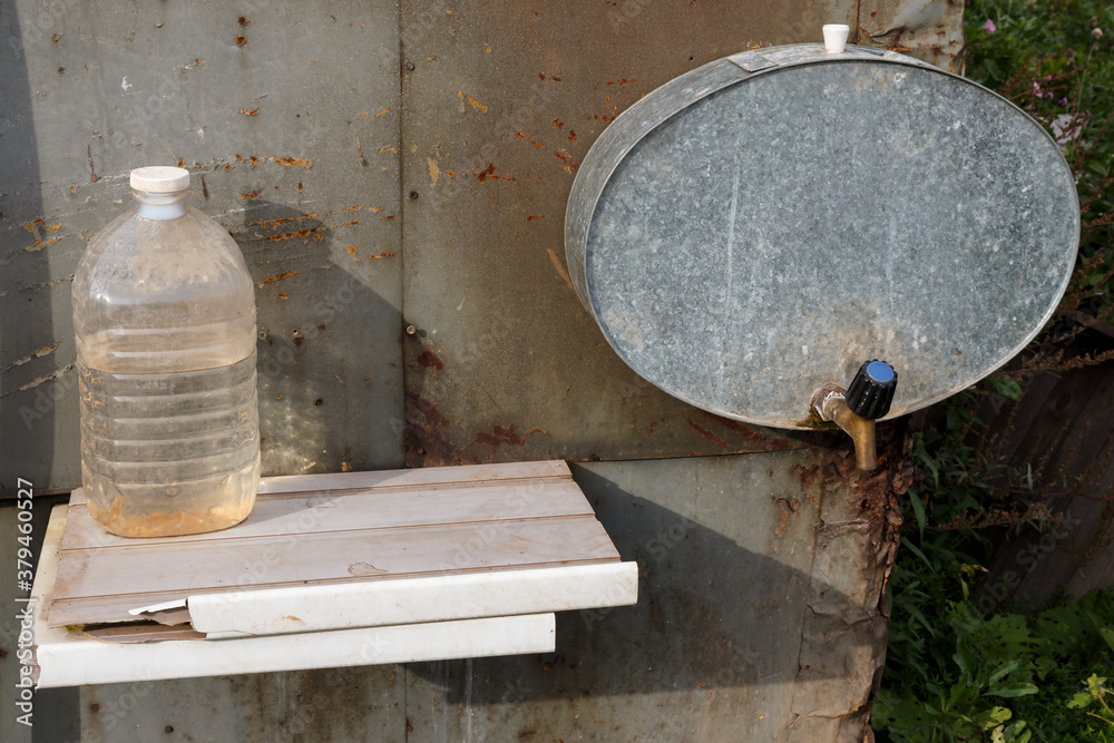 Country washbasin. An iron tank with a tap hangs on the wall. Washbasin in the garden and a bottle of water.