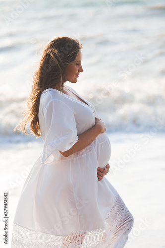 Beautiful elegant pregnant woman 25-26 year old wearing white trendy dress holding tummy with hands posing over sea shore background close up. Motherhood. Maternity.