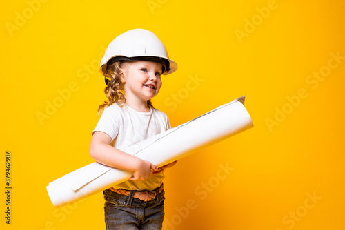 Cute little girl engineer with helmet holding bluepring on yellow background