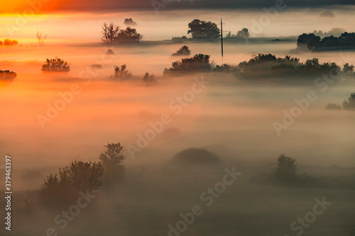 Beautiful panoramic landscape with river valley covered by thick fog in autumn in the early morning. Sunrise. Sun rays shine through the thick fog. Electric power transmission line in fog.
