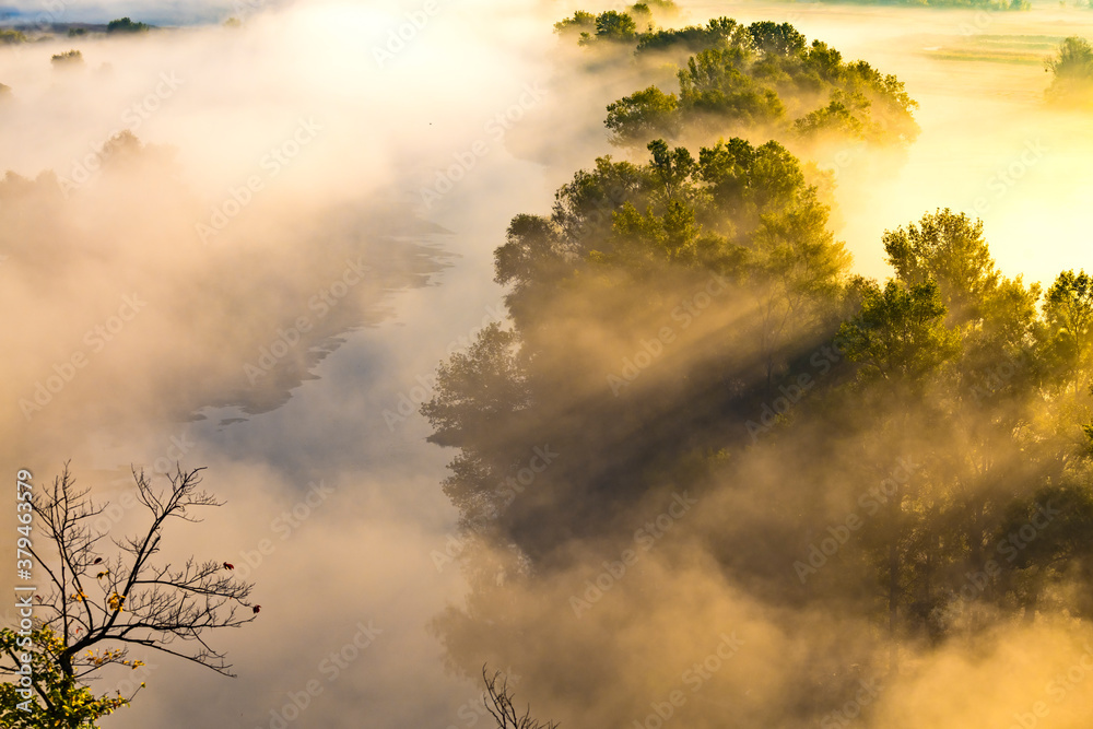 Beautiful panoramic landscape with river valley covered by thick fog in autumn in the early morning. Sunrise. Sun rays shine through the thick fog.