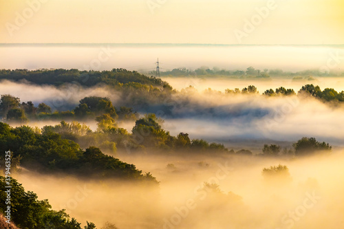 Beautiful panoramic landscape with river valley covered by thick fog in autumn in the early morning. Sunrise. Sun rays shine through the thick fog. Electric power transmission line in fog.