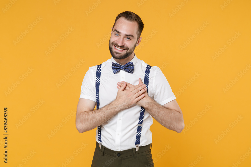 Smiling pleasant handsome young bearded man 20s wearing white shirt bow-tie suspender posing holding hands folded on heart looking camera isolated on bright yellow color background, studio portrait. Smiling pleasant handsome young bearded man 20s wearing white shirt bow-tie suspender posing holding hands folded on heart looking camera isolated on bright yellow color background, studio portrait.