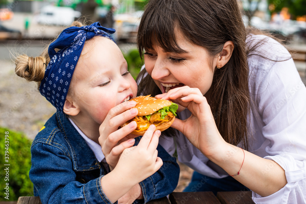Young mother and little daughter ear burger together in outdoors cafe