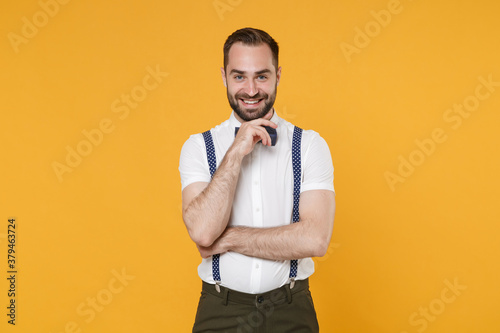 Smiling handsome young bearded man 20s wearing white shirt bow-tie suspender posing standing put hand prop up on chin looking camera isolated on bright yellow color wall background, studio portrait. © ViDi Studio