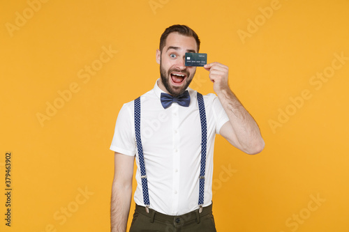 Excited cheerful funny young bearded man 20s wearing white shirt bow-tie suspender posing standing covering eye with credit bank card isolated on bright yellow color wall background studio portrait.