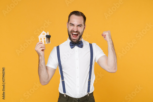 Screaming young bearded man 20s wearing white shirt bow-tie suspender posing standing hold in hand house bunch of keys doing winner gesture isolated on bright yellow color background studio portrait.