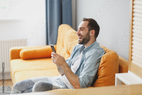 Side view of cheerful funny young bearded man wearing casual white t-shirt blue shirt using mobile cell phone typing sms message sitting on couch resting relaxing spending time in living room at home.