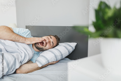 Funny young bearded man wearing basic blue t-shirt sleeping yawning covering mouth with hand lying in bed with striped sheet pillow blanket resting relaxing spending time in bedroom at home.
