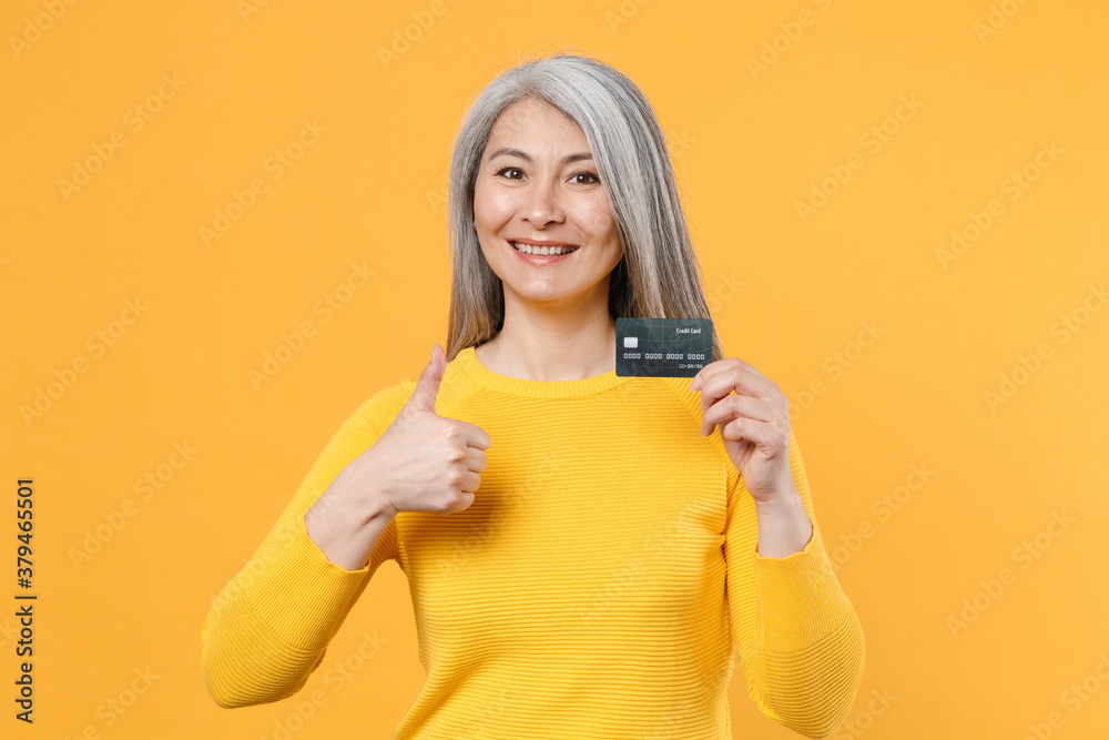 Smiling beautiful gray-haired asian woman wearing casual clothes standing holding in hand credit bank card showing thumb up looking camera isolated on bright yellow colour background, studio portrait. Smiling beautiful gray-haired asian woman wearing casual clothes standing holding in hand credit bank card showing thumb up looking camera isolated on bright yellow colour background, studio portrait.