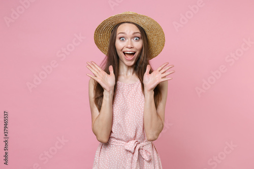 Shocked excited surprised attractive young brunette woman 20s in pink summer dotted dress hat posing keeping mouth open saying wow spreading hands isolated on pastel pink background studio portrait.