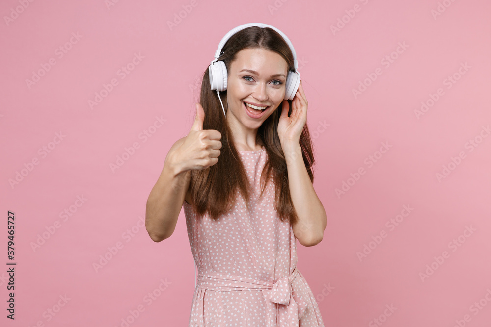 Cheerful beautiful young brunette woman 20s in pink summer dotted dress posing listening music with headphones showing thumb up looking camera isolated on pastel pink color background studio portrait. Cheerful beautiful young brunette woman 20s in pink summer dotted dress posing listening music with headphones showing thumb up looking camera isolated on pastel pink color background studio portrait.