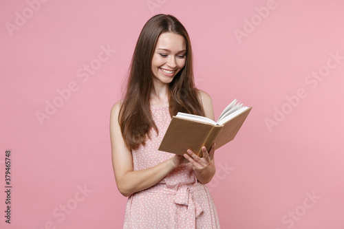 Smiling funny pretty attractive beautiful young brunette woman 20s wearing pink summer dotted dress posing holding in hands reading book isolated on pastel pink color wall background studio portrait.