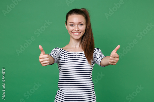 Smiling cheerful funny beautiful young brunette woman 20s wearing striped casual clothes posing standing showing thumbs up looking camera isolated on green color wall background, studio portrait.