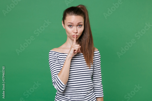 Secret pretty young brunette woman 20s wearing striped casual clothes posing saying hush be quiet with finger on lips shhh gesture looking camera isolated on green color background, studio portrait.