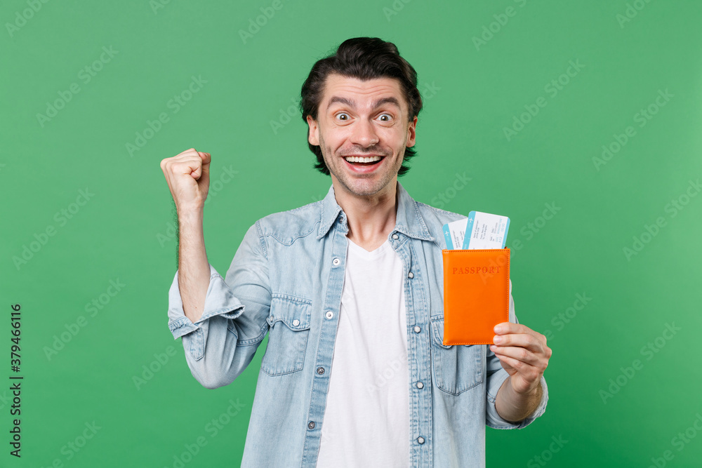 Happy young traveler tourist man 20s in casual clothes white t-shirt denim shirt hold passport ticket boarding pass doing winner gesture looking camera isolated on green background studio portrait. Happy young traveler tourist man 20s in casual clothes white t-shirt denim shirt hold passport ticket boarding pass doing winner gesture looking camera isolated on green background studio portrait.