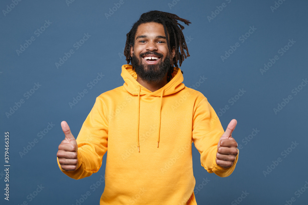 Cheerful young african american man guy wearing casual yellow streetwear hoodie posing isolated on blue wall background studio portrait. People sincere emotions lifestyle concept. Showing thumbs up. Cheerful young african american man guy wearing casual yellow streetwear hoodie posing isolated on blue wall background studio portrait. People sincere emotions lifestyle concept. Showing thumbs up.