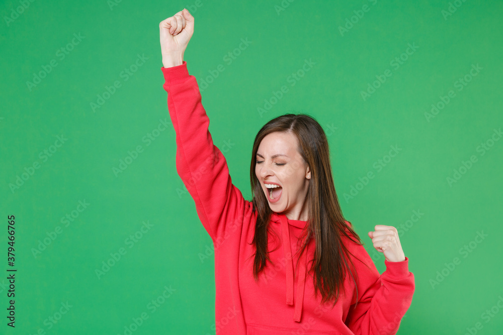 Overjoyed happy screaming young brunette woman 20s in red casual streetwear hoodie posing clenching fists doing winner gesture keeping eyes closed isolated on green color background studio portrait.