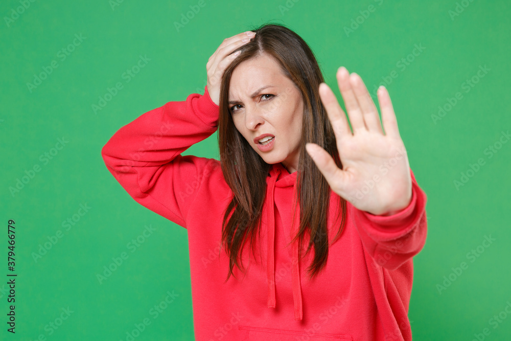 Displeased tired young brunette woman 20s in red casual streetwear hoodie posing showing stop gesture with palm put hand on head looking camera isolated on green color background studio portrait. Displeased tired young brunette woman 20s in red casual streetwear hoodie posing showing stop gesture with palm put hand on head looking camera isolated on green color background studio portrait.