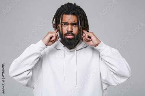 Upset young african american man guy with dreadlocks 20s wearing white casual streetwear hoodie posing looking camera covering ears with fingers isolated on grey color wall background studio portrait.