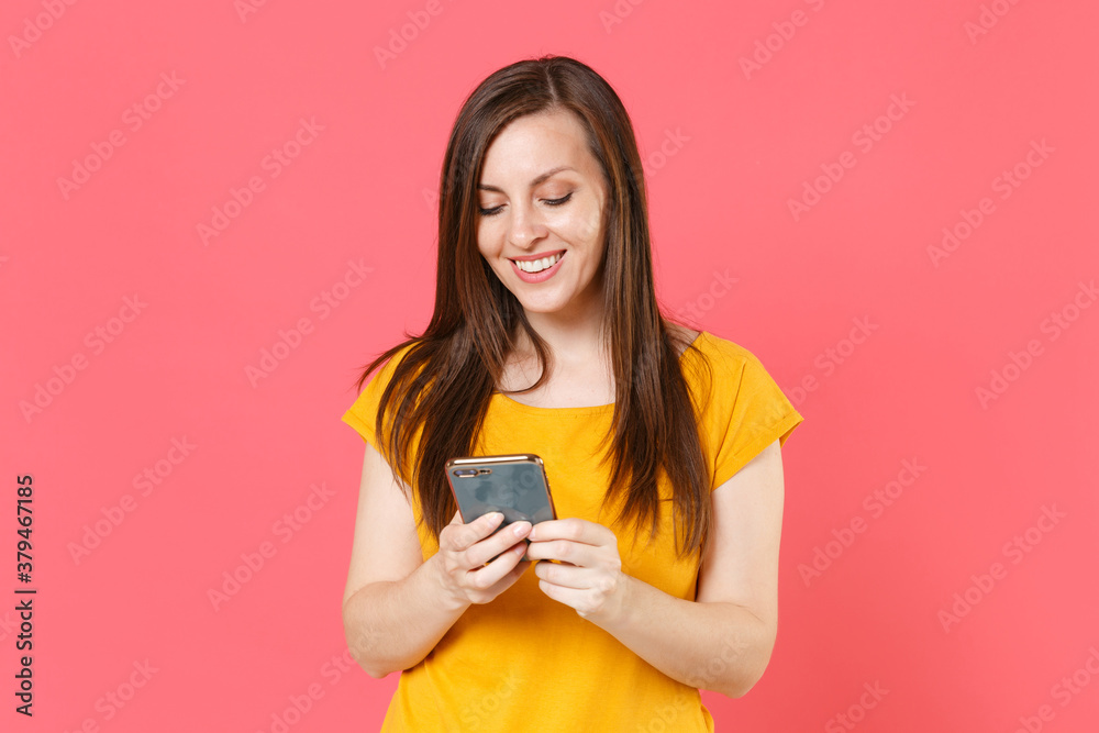 Smiling beautiful attractive young brunette woman 20s wearing yellow casual t-shirt posing standing using mobile cell phone typing sms message isolated on pink color wall background studio portrait. Smiling beautiful attractive young brunette woman 20s wearing yellow casual t-shirt posing standing using mobile cell phone typing sms message isolated on pink color wall background studio portrait.