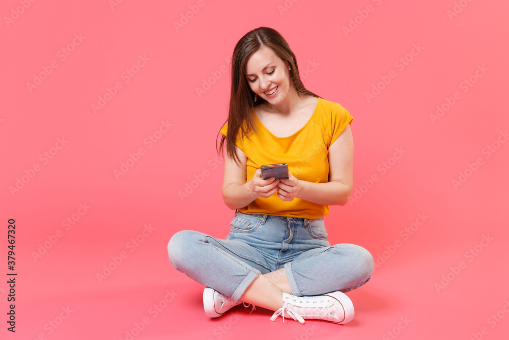 Full length portrait of smiling beautiful attractive young brunette woman 20s in yellow t-shirt sit on floor using mobile cell phone typing sms message isolated on pink color wall background studio. Full length portrait of smiling beautiful attractive young brunette woman 20s in yellow t-shirt sit on floor using mobile cell phone typing sms message isolated on pink color wall background studio.