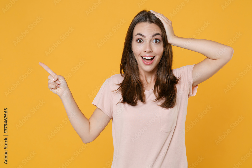 Excited young brunette woman 20s in pastel pink casual t-shirt standing posing point index finger aside up on mock up copy space put hand on head isolated on yellow color background studio portrait. Excited young brunette woman 20s in pastel pink casual t-shirt standing posing point index finger aside up on mock up copy space put hand on head isolated on yellow color background studio portrait.
