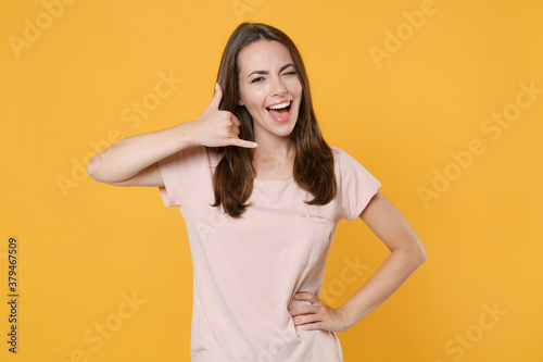 Blinking funny young brunette woman 20s wearing pastel pink casual t-shirt standing doing phone gesture like says call me back looking camera isolated on yellow color wall background studio portrait.