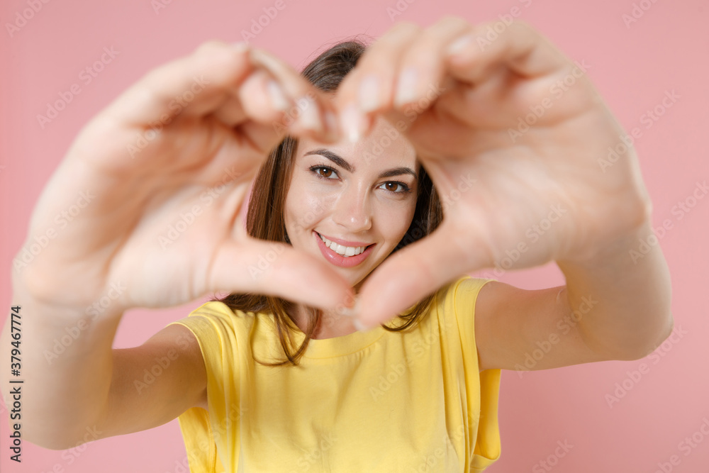 Smiling young brunette woman 20s wearing yellow casual t-shirt posing standing showing shape heart with hands, heart-shape sign looking camera isolated on pastel pink color background studio portrait. Smiling young brunette woman 20s wearing yellow casual t-shirt posing standing showing shape heart with hands, heart-shape sign looking camera isolated on pastel pink color background studio portrait.