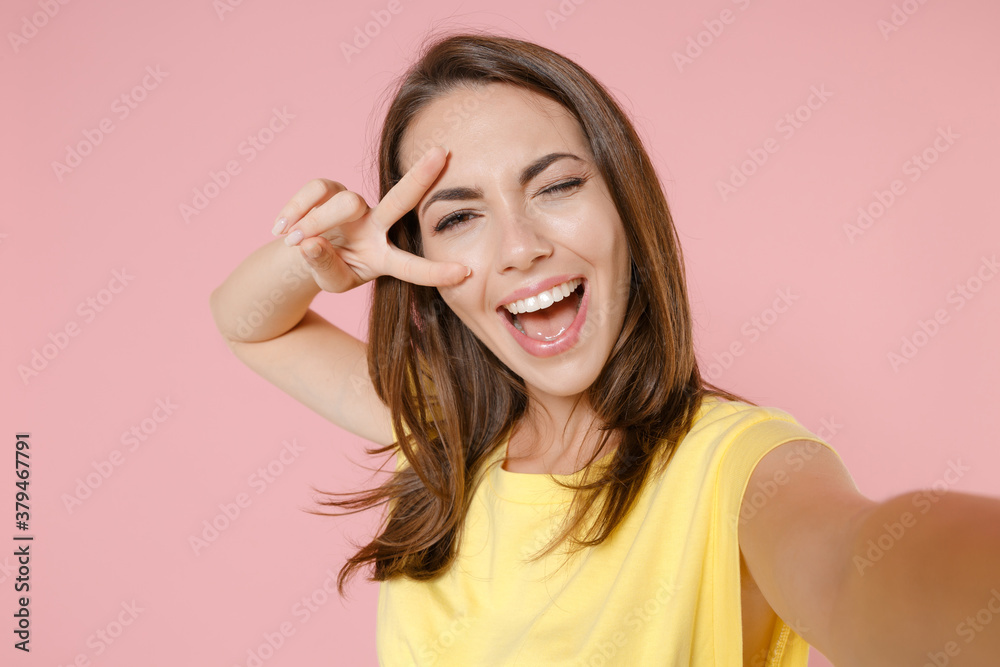 Close up of blinking young brunette woman 20s in yellow casual t-shirt posing standing doing selfie shot on mobile phone showing victory sign isolated on pastel pink color background studio portrait. Close up of blinking young brunette woman 20s in yellow casual t-shirt posing standing doing selfie shot on mobile phone showing victory sign isolated on pastel pink color background studio portrait.