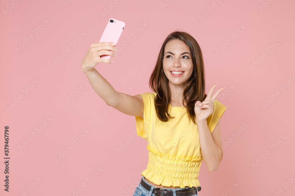 Smiling young brunette woman 20s wearing yellow casual t-shirt posing standing doing selfie shot on mobile phone showing victory sign isolated on pastel pink color wall background studio portrait. Smiling young brunette woman 20s wearing yellow casual t-shirt posing standing doing selfie shot on mobile phone showing victory sign isolated on pastel pink color wall background studio portrait.