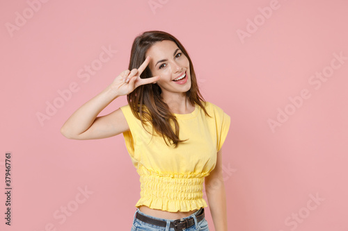 Cheerful funny smiling young brunette woman 20s wearing yellow casual t-shirt posing standing showing victory sign looking camera isolated on pastel pink color wall background studio portrait.