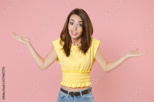 Confused puzzled perplexed young brunette woman 20s wearing yellow casual t-shirt posing standing spreading hands looking camera isolated on pastel pink color wall background studio portrait.