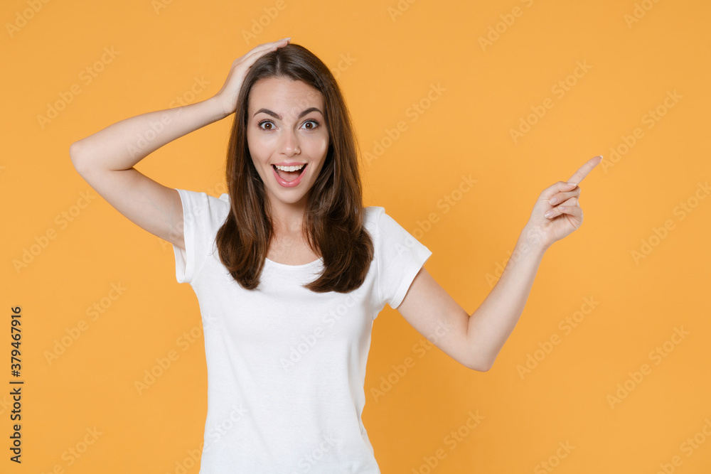 Excited young brunette woman 20s in white blank empty design casual t-shirt pointing index finger aside up on mock up copy space put hand on head isolated on yellow color background studio portrait. Excited young brunette woman 20s in white blank empty design casual t-shirt pointing index finger aside up on mock up copy space put hand on head isolated on yellow color background studio portrait.