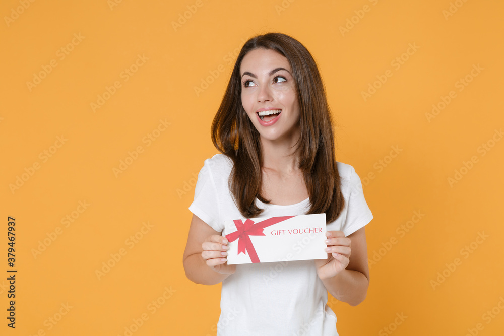 Cheerful funny smiling young brunette woman 20s wearing white blank design casual t-shirt posing holding in hands gift certificate looking aside isolated on yellow color background studio portrait.