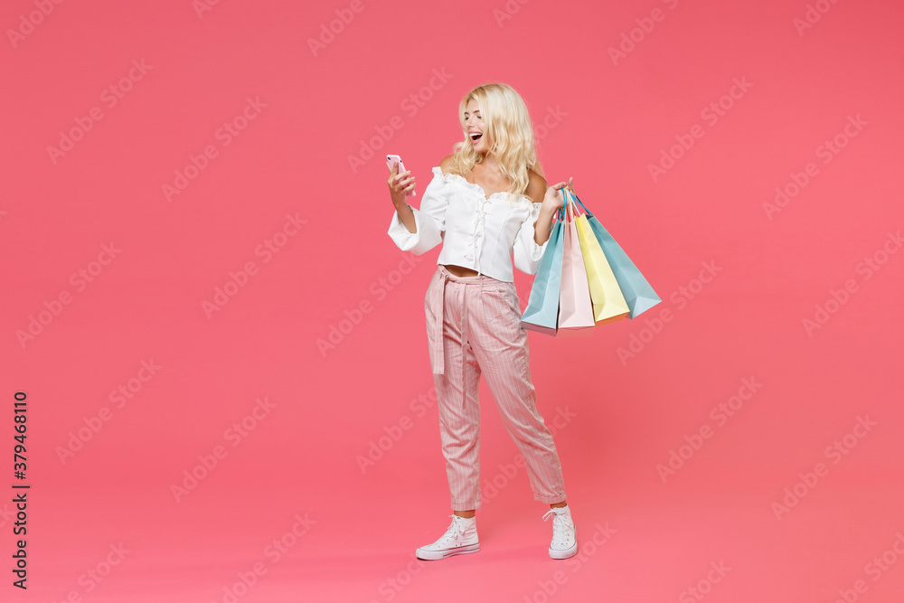 Full length portrait of funny young blonde woman 20s in white casual clothes hold package bag with purchases after shopping using mobile cell phone isolated on bright pink colour background studio. Full length portrait of funny young blonde woman 20s in white casual clothes hold package bag with purchases after shopping using mobile cell phone isolated on bright pink colour background studio.