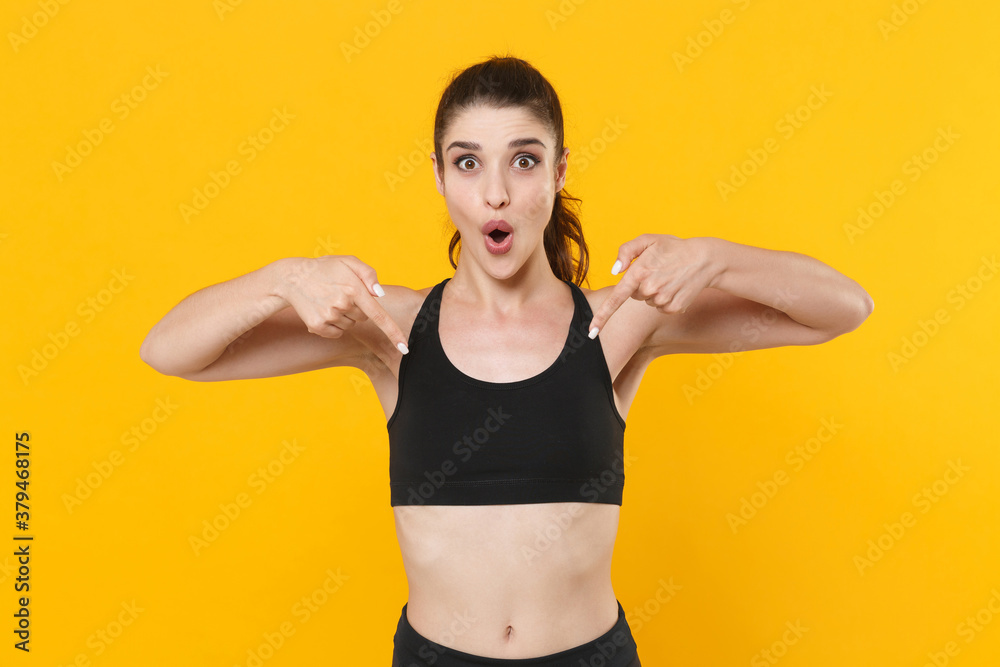 Shocked young fitness sporty woman 20s wearing black sportswear posing training working out pointing index fingers down on mock up copy space isolated on yellow color wall background studio portrait. Shocked young fitness sporty woman 20s wearing black sportswear posing training working out pointing index fingers down on mock up copy space isolated on yellow color wall background studio portrait.