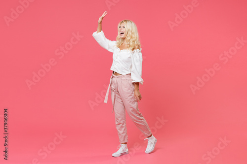 Full length portrait of cheerful young blonde woman 20s wearing white casual clothes waving and greeting with hand as notices someone looking aside isolated on bright pink colour background in studio.