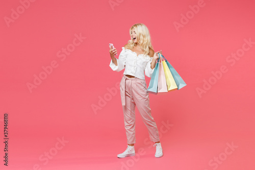 Full length portrait of funny young blonde woman 20s in white casual clothes hold package bag with purchases after shopping using mobile cell phone isolated on bright pink colour background studio.