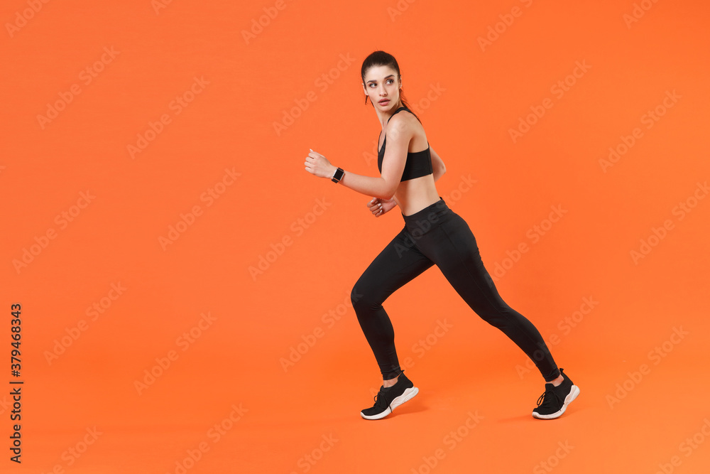 Full length side view portrait of beautiful strong young fitness sporty woman 20s wearing black sportswear training working out running looking aside isolated on orange color wall background studio.